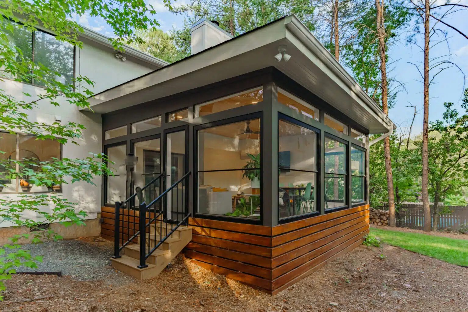 Exterior view of a modern screened-in porch addition with a natural wood-slat base, black-framed windows, and integrated stairs leading into a wooded backyard.