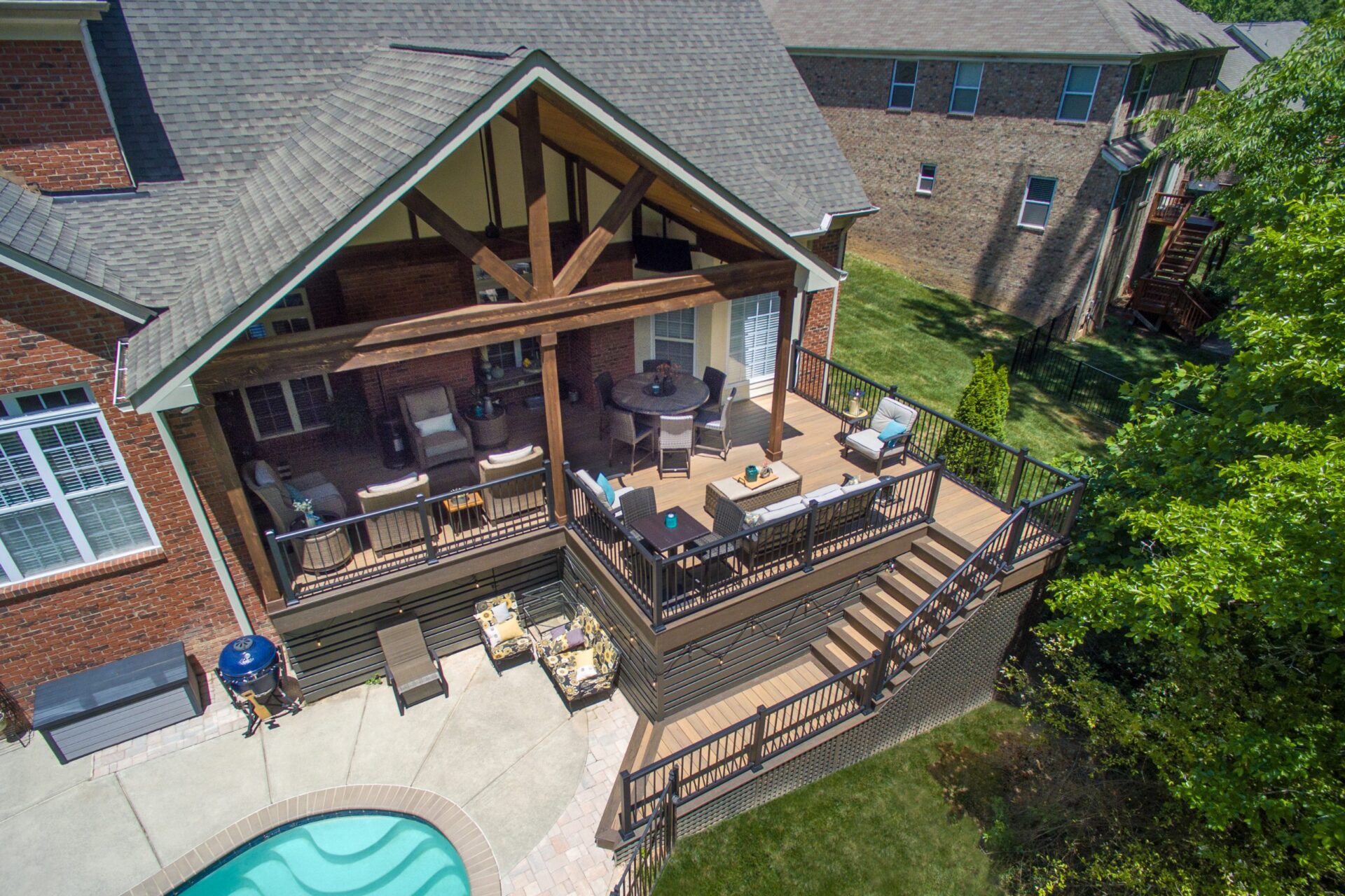 An aerial top-down view of a luxury wood-framed patio cover protecting an outdoor living space with a sofa, fireplace, and deck overlooking a swimming pool.