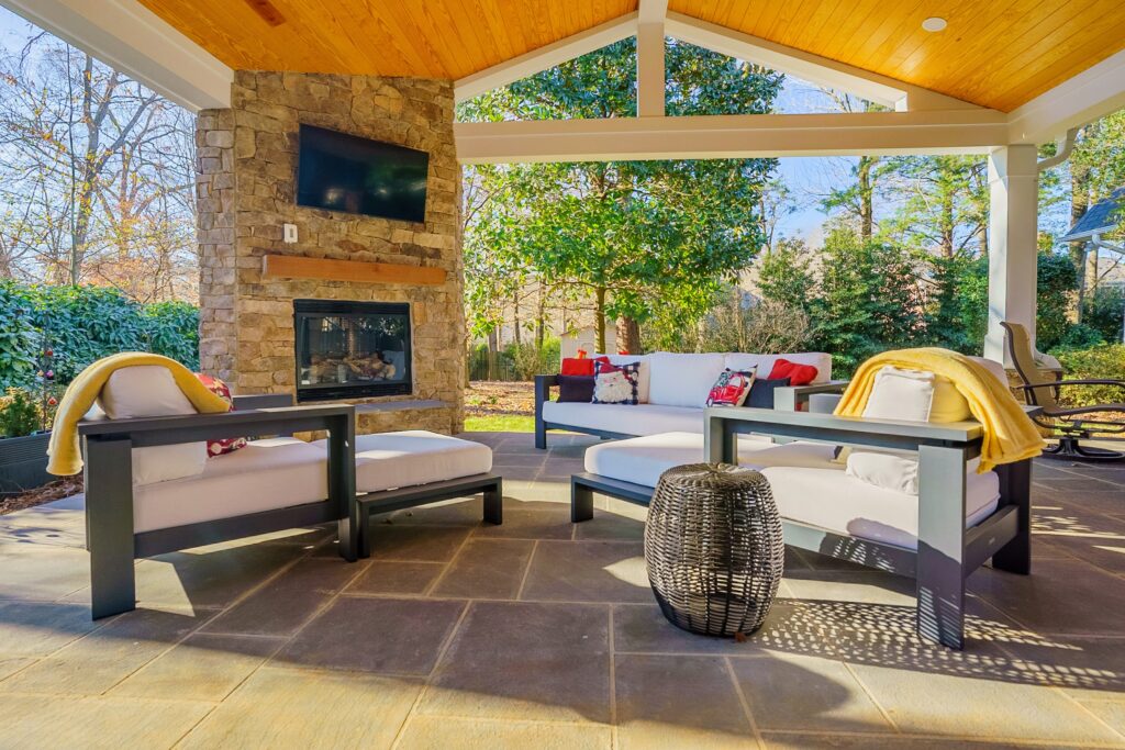 Luxury covered patio featuring a rustic stone fireplace with mounted TV, wood ceiling, and comfortable outdoor lounge chairs.