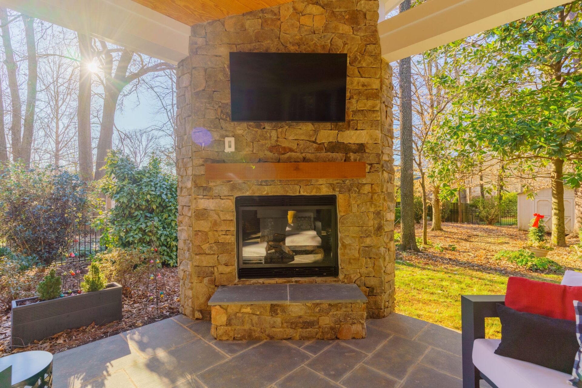 Outdoor living area featuring a stacked stone fireplace with mounted TV, wood mantel, and dark tile patio, custom built by Carolina Decks in the Marvin area.