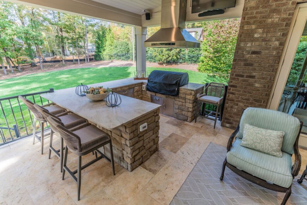Spacious covered patio features a stone outdoor kitchen, long bar, stainless hood, and travertine floor, custom built by Carolina Decks in the Charlotte, NC area.