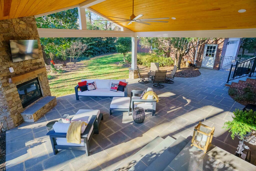 Spacious covered paver patio featuring a stone fireplace, wood ceiling, and comfortable outdoor furniture custom built by Carolina Decks in the Soutpark area.