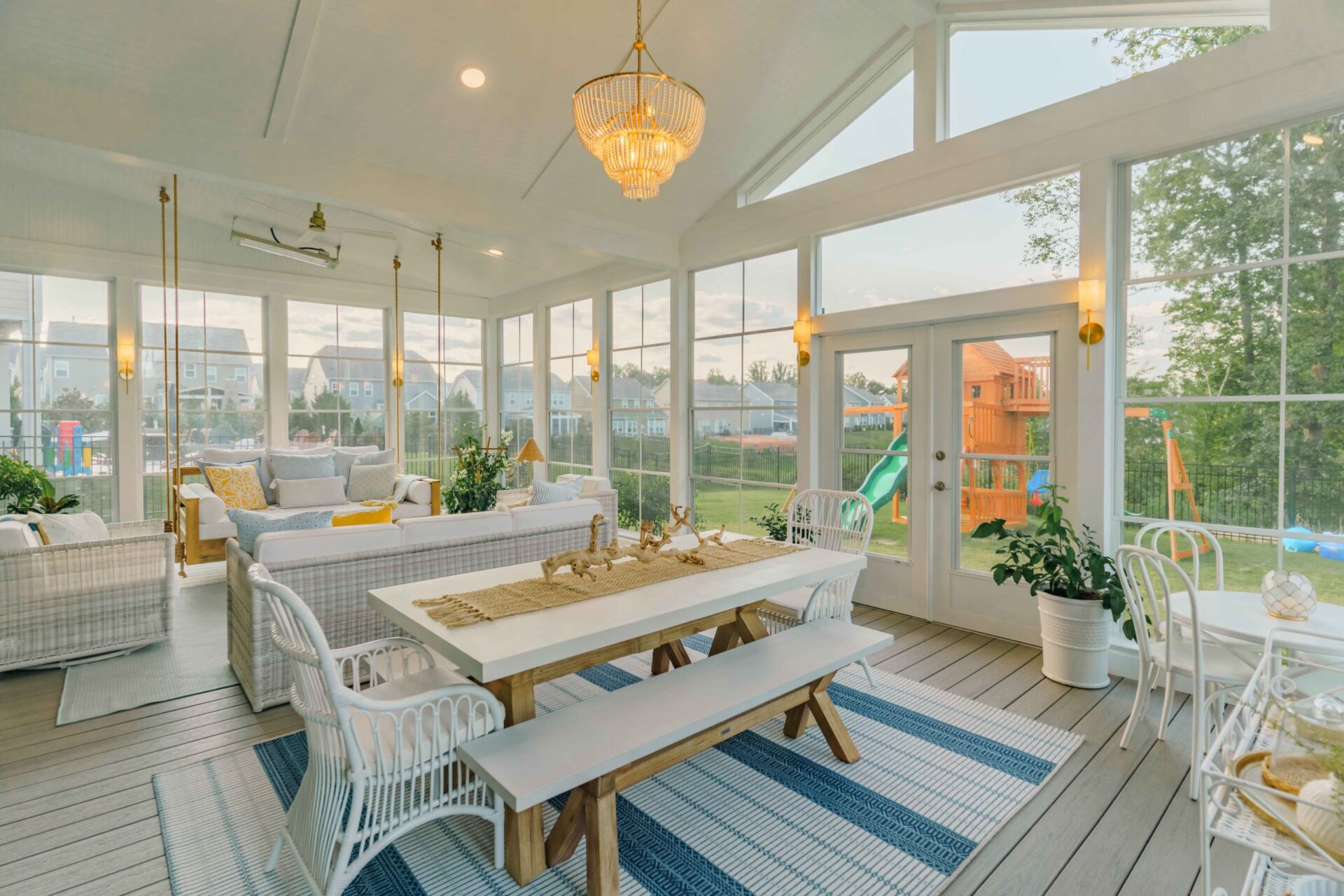 Wide-angle view of a beautifully styled, upscale screened porch in the Millbridge neighborhood, highlighting the wood dining table, white wicker furniture, crystal chandelier, and a view of the backyard playset outside.
