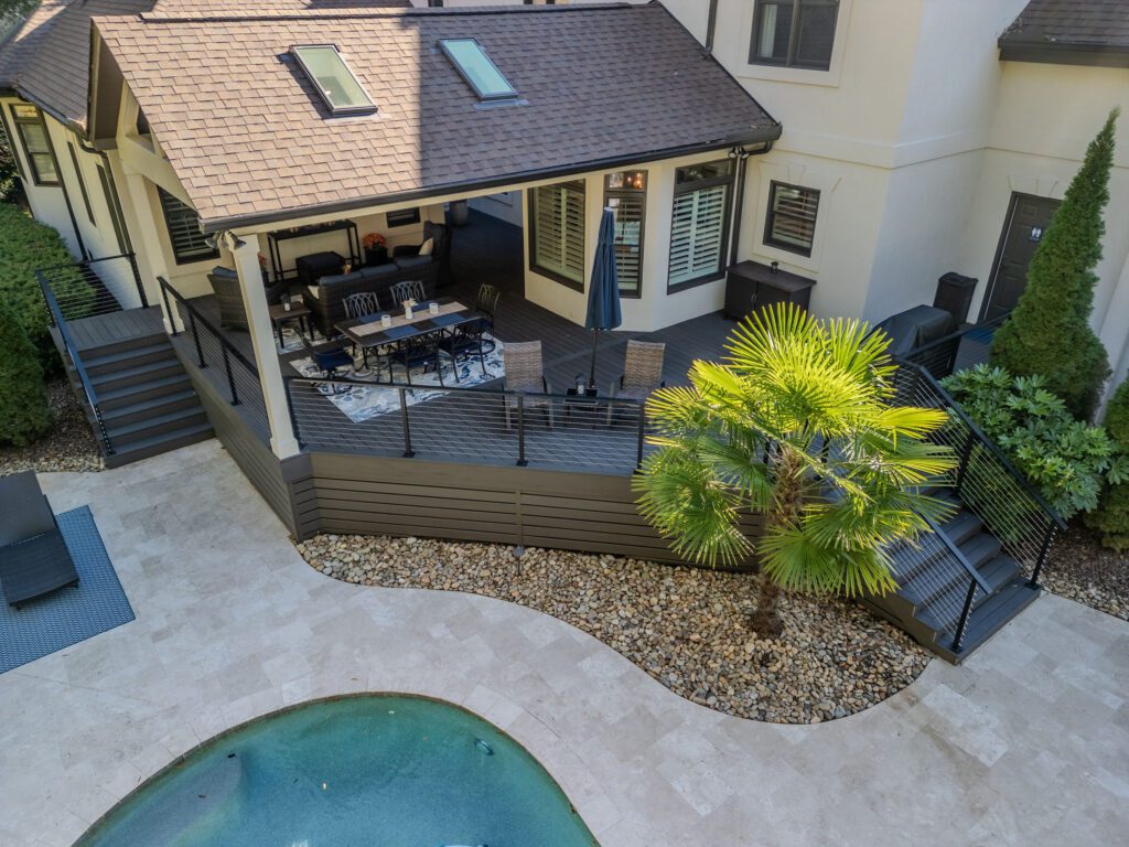 An elegant covered composite deck with cable railings and steps leads to a travertine paver patio and pool, custom built by Carolina Decks in the Providence Country Club neighborhood.