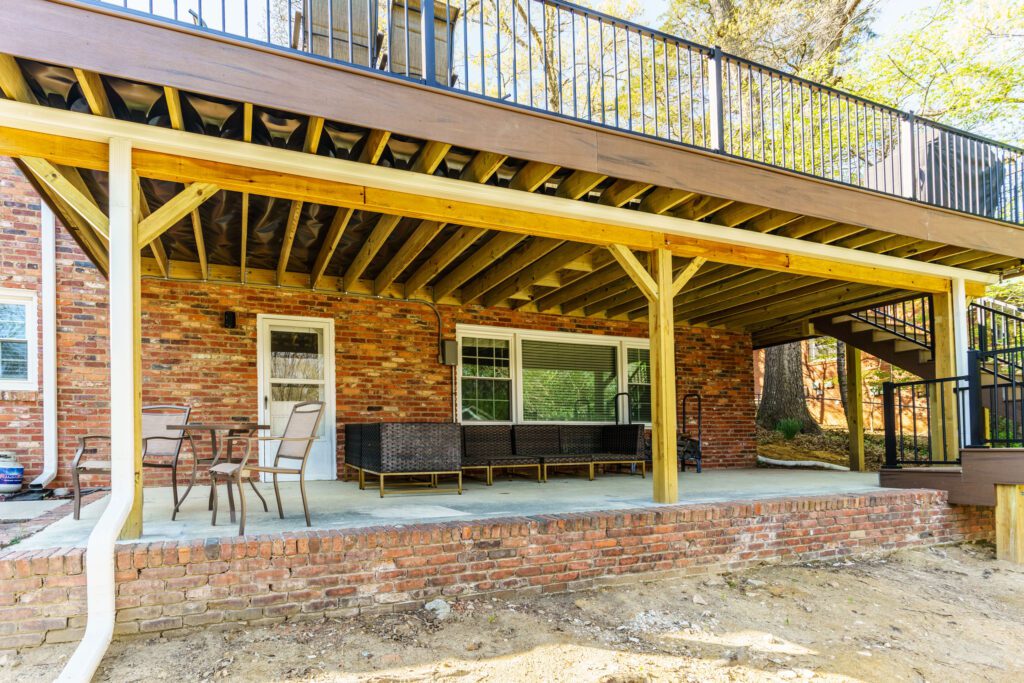 A ground-level brick patio underneath an elevated deck in Charlotte's Stonehaven neighborhood, showing support beams, outdoor furniture, and a drainage system ceiling.