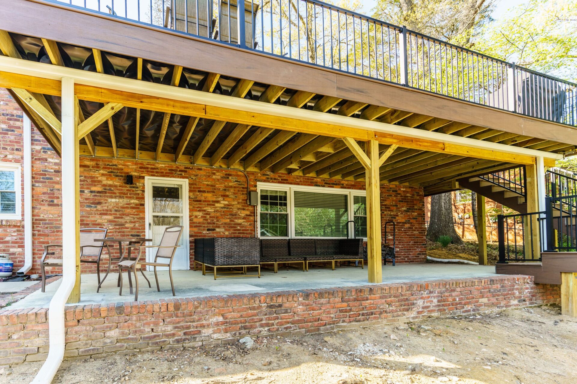A ground-level brick patio underneath an elevated deck in Charlotte's Stonehaven neighborhood, showing support beams, outdoor furniture, and a drainage system ceiling.