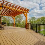 Large tan composite deck with a custom timber pergola, black metal railings, and stone posts, custom built by Carolina Decks in the Lake Norman, NC area.