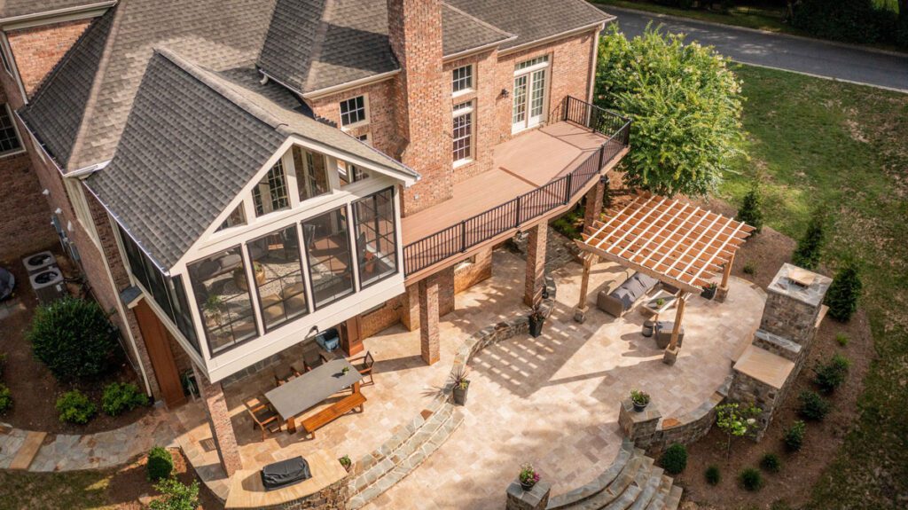 Aerial view of a luxury multi-level outdoor living project in Piper Glen, Charlotte, featuring a TimberTech deck, an enclosed Eze-Breeze sunroom, a large stone patio with an outdoor fireplace, a custom wood pergola, and an integrated dining area.