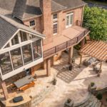 Aerial view of a luxury multi-level outdoor living project in Piper Glen, Charlotte, featuring a TimberTech deck, an enclosed Eze-Breeze sunroom, a large stone patio with an outdoor fireplace, a custom wood pergola, and an integrated dining area.