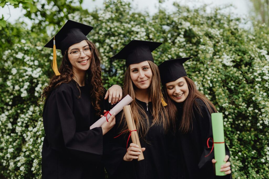 Three happy graduates in caps and gowns standing in a green spring backyard in Charlotte, NC.