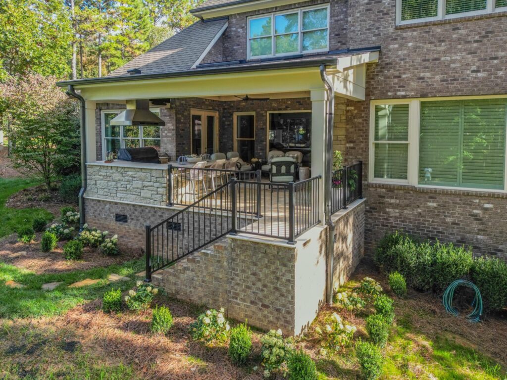 Exterior elevated view of a custom covered porch addition with brick foundation, stone kitchen wall, and railing built by Carolina Decks in Mint Hill NC.