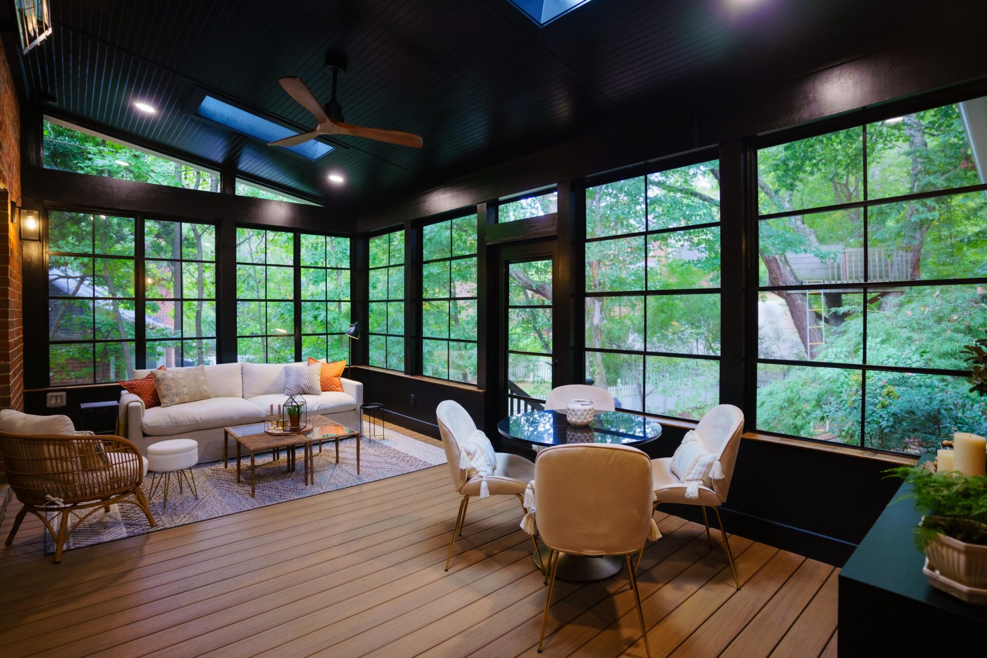 Interior hero shot of a luxury screened-in porch featuring a dark vaulted ceiling with skylights, a ceiling fan, large grid windows looking out to trees, composite flooring, and modern living and dining furniture.