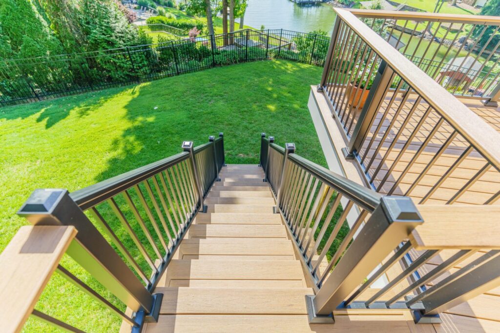 Looking down a set of custom deck stairs built with TimberTech Coconut Husk composite decking and black aluminum railing in a Tega Cay neighborhood backyard overlooking a lake.