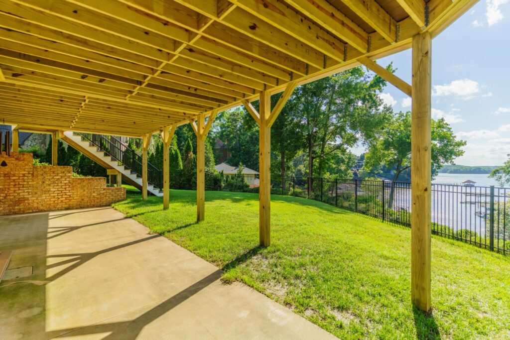 The under-deck structural wood framing and concrete patio of a two-story deck build in Tega Cay, featuring views of the lawn, black aluminum perimeter fence, and lake.