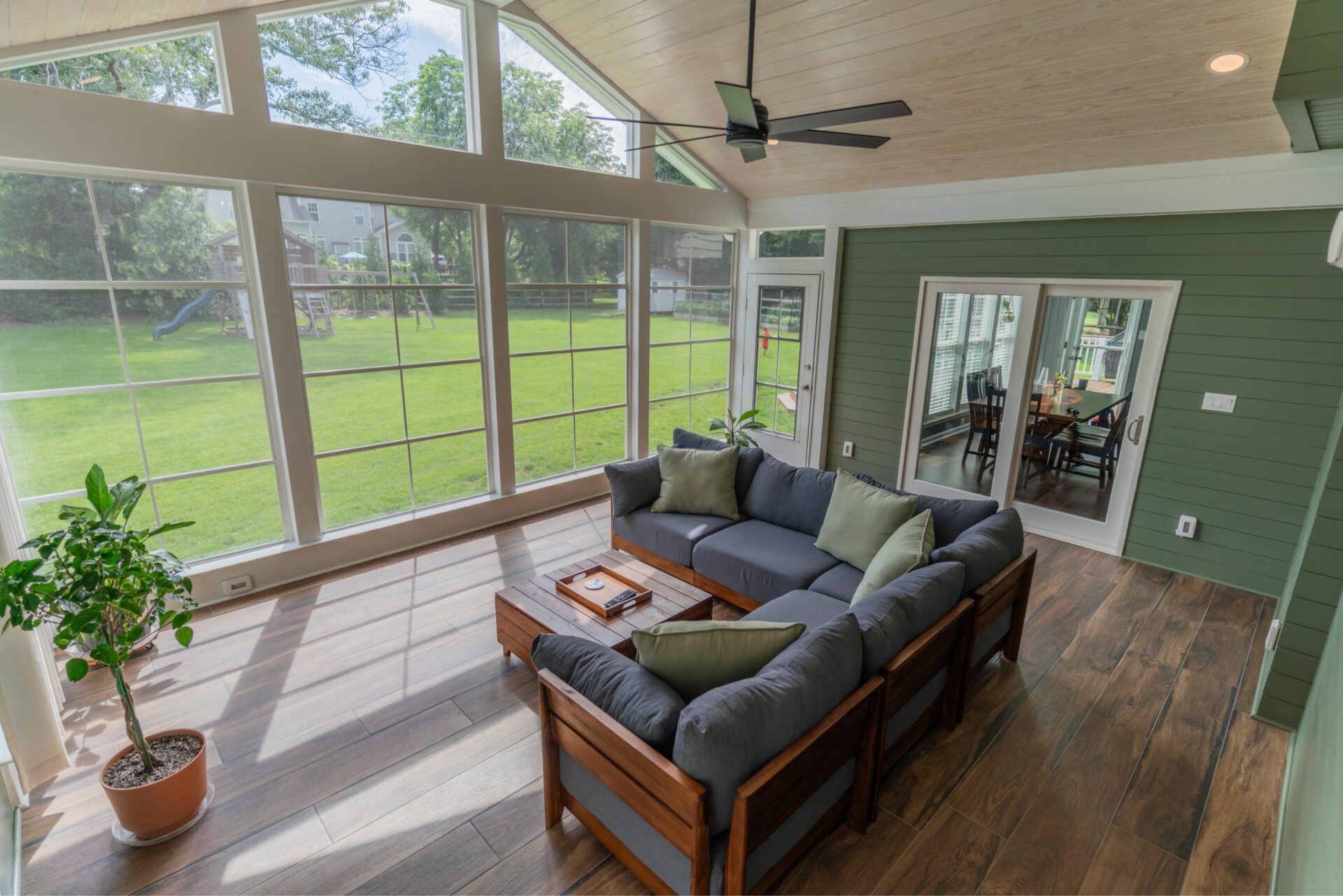 Interior overview of an enclosed Ezebreeze porch featuring Tobacco Cask wood plank matte porcelain tile floors, a vaulted ceiling, a grey sectional sofa, and a green shiplap wall with sliding glass doors.