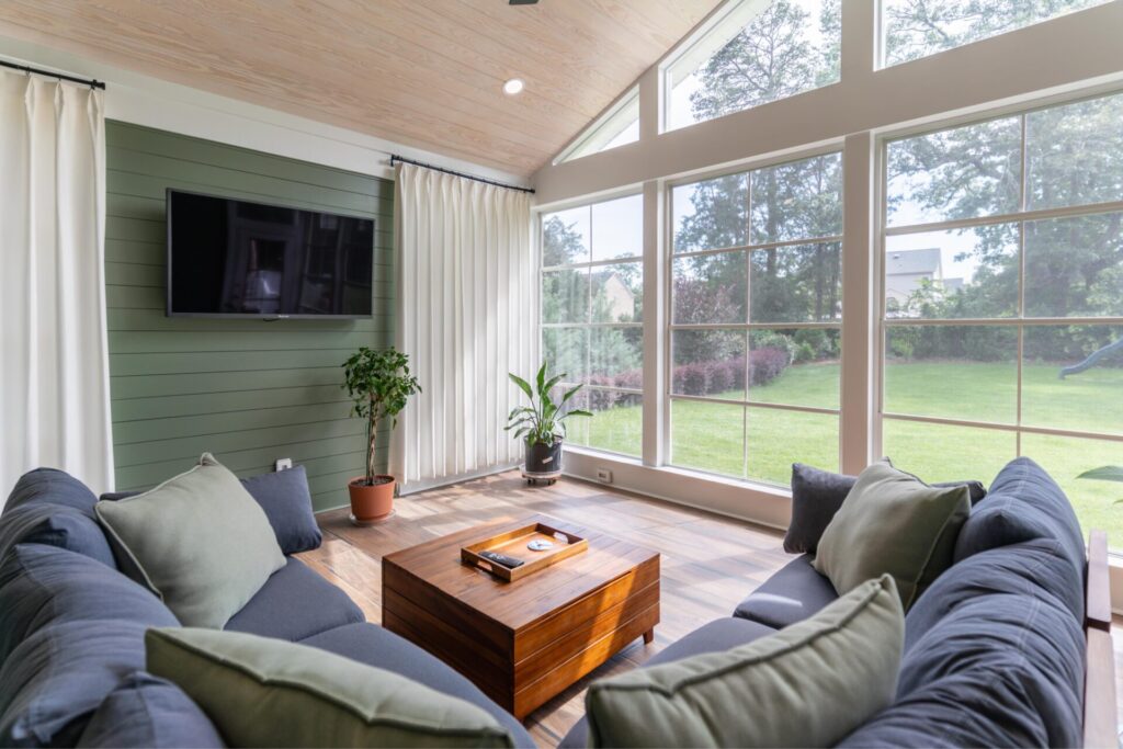 Cozy enclosed porch living room featuring a green shiplap feature wall with a mounted TV, comfortable grey seating, and large Ezebreeze window panels letting in natural light.