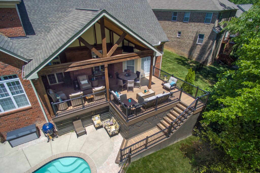 An aerial top-down view of a luxury wood-framed patio cover protecting an outdoor living space with a sofa, fireplace, and deck overlooking a swimming pool.