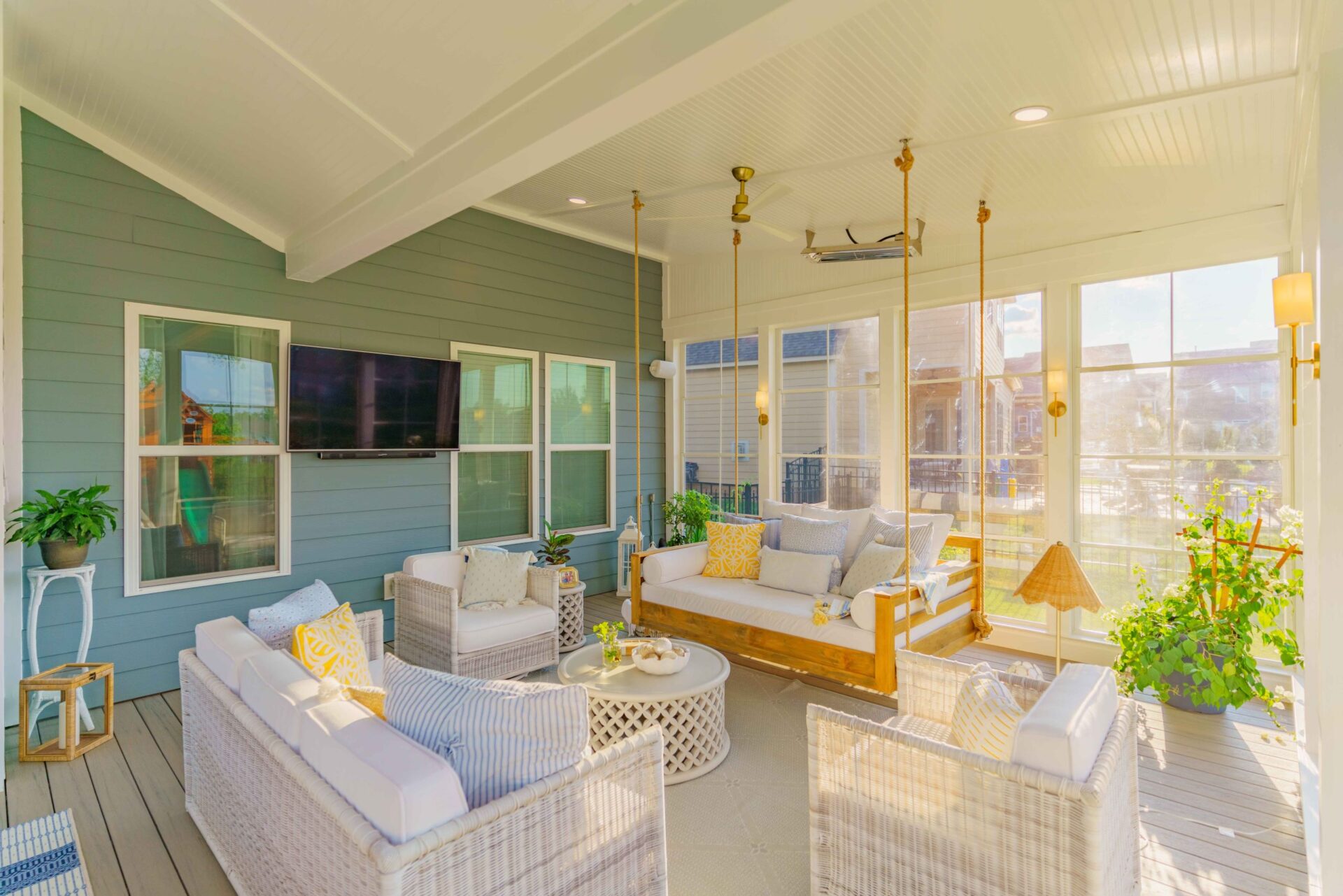 Interior view of an upscale outdoor living room on a screened porch, highlighting a wall-mounted TV, cozy white wicker seating, a hanging rope daybed swing, and modern wall sconce lighting.