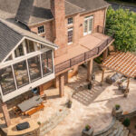 Aerial view of a luxury multi-level outdoor living project in Piper Glen, Charlotte, featuring a TimberTech deck, an enclosed Eze-Breeze sunroom, a large stone patio with an outdoor fireplace, a custom wood pergola, and an integrated dining area.