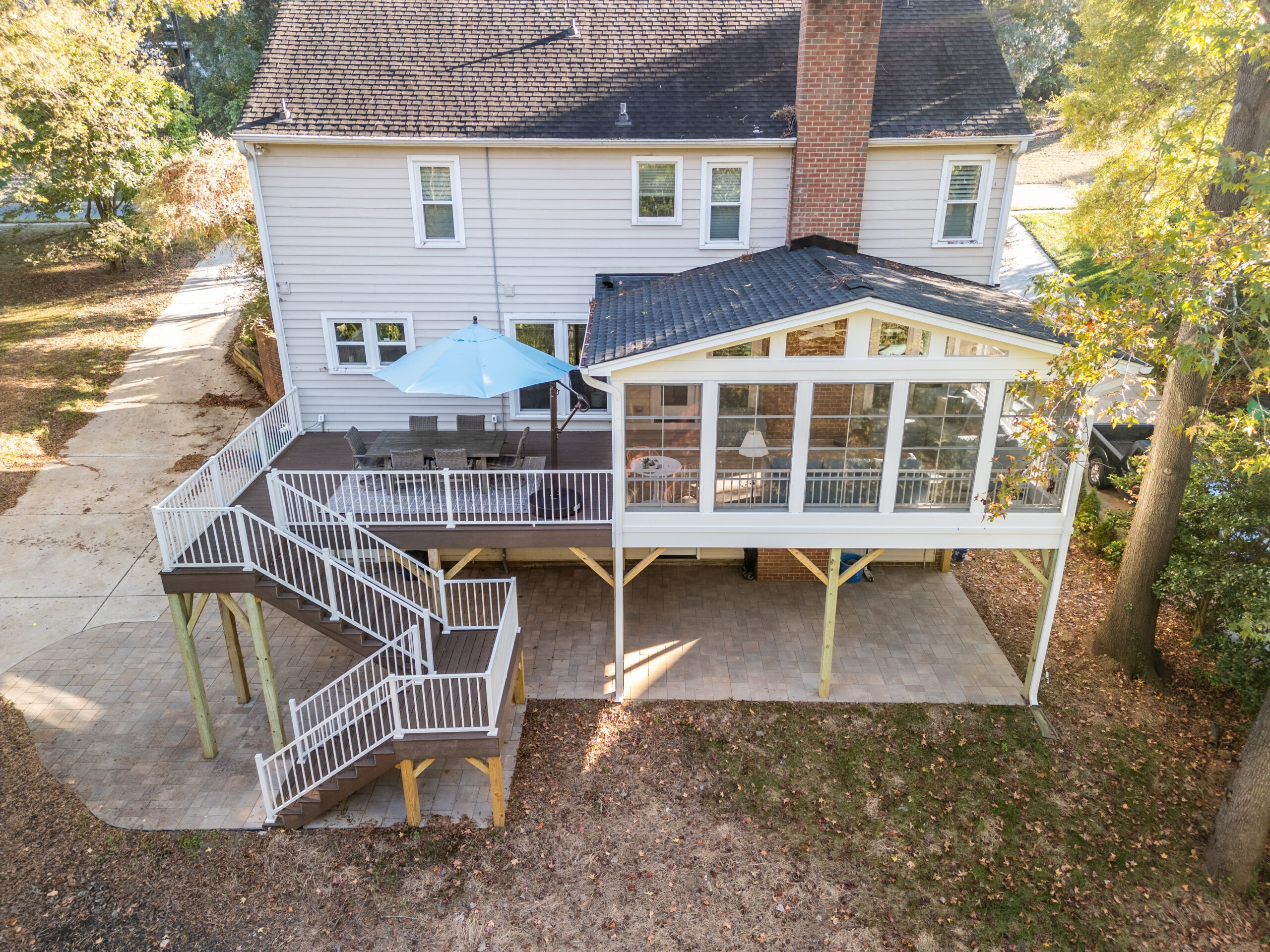 Aerial view of a two-story outdoor living space in the Carmel neighborhood of Charlotte, NC featuring an Eze-Breeze 3 season porch with gabled roof, brown TimberTech composite decking, white aluminum railings, and a lower-level paver patio.